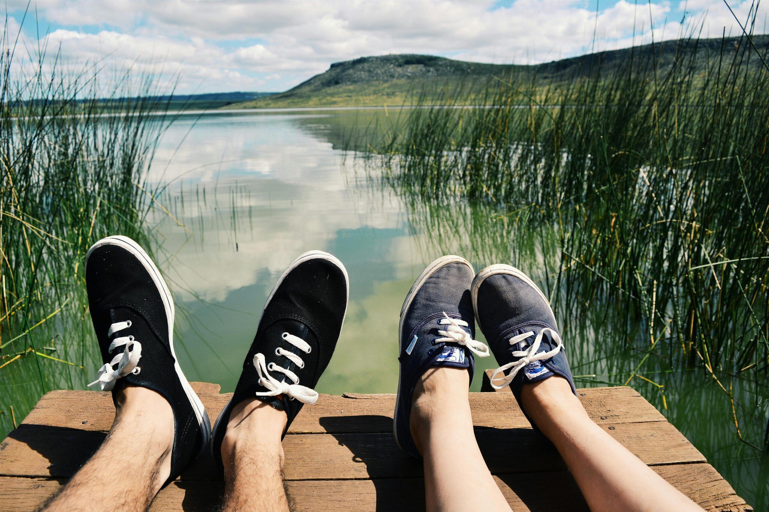 Two people relaxing by a lake with reeds and hills in the background on a sunny day.